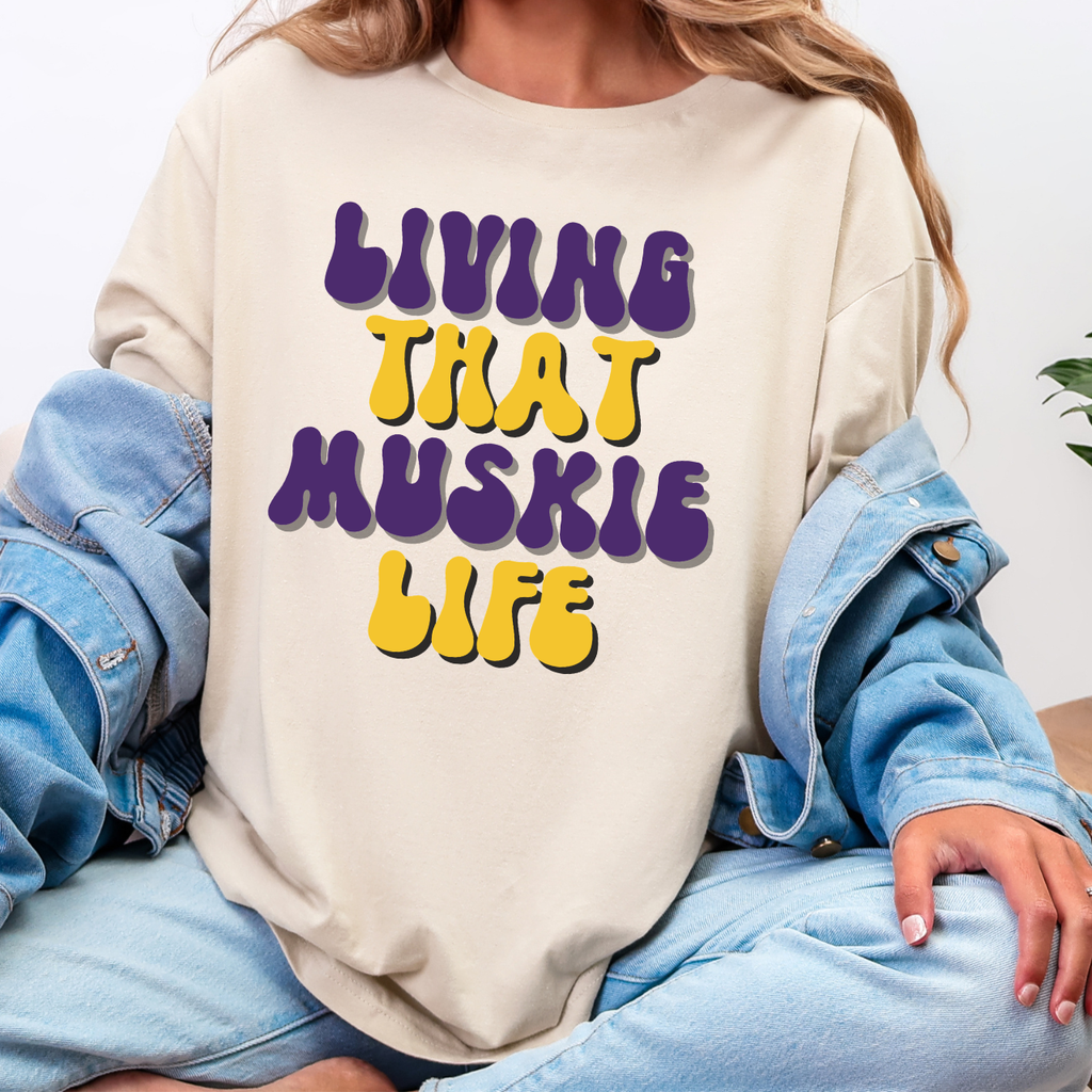 Person wearing a beige sweatshirt with 'Living That Muskie Life' text in purple and yellow.