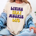 Person wearing a beige sweatshirt with 'Living That Muskie Life' text in purple and yellow.