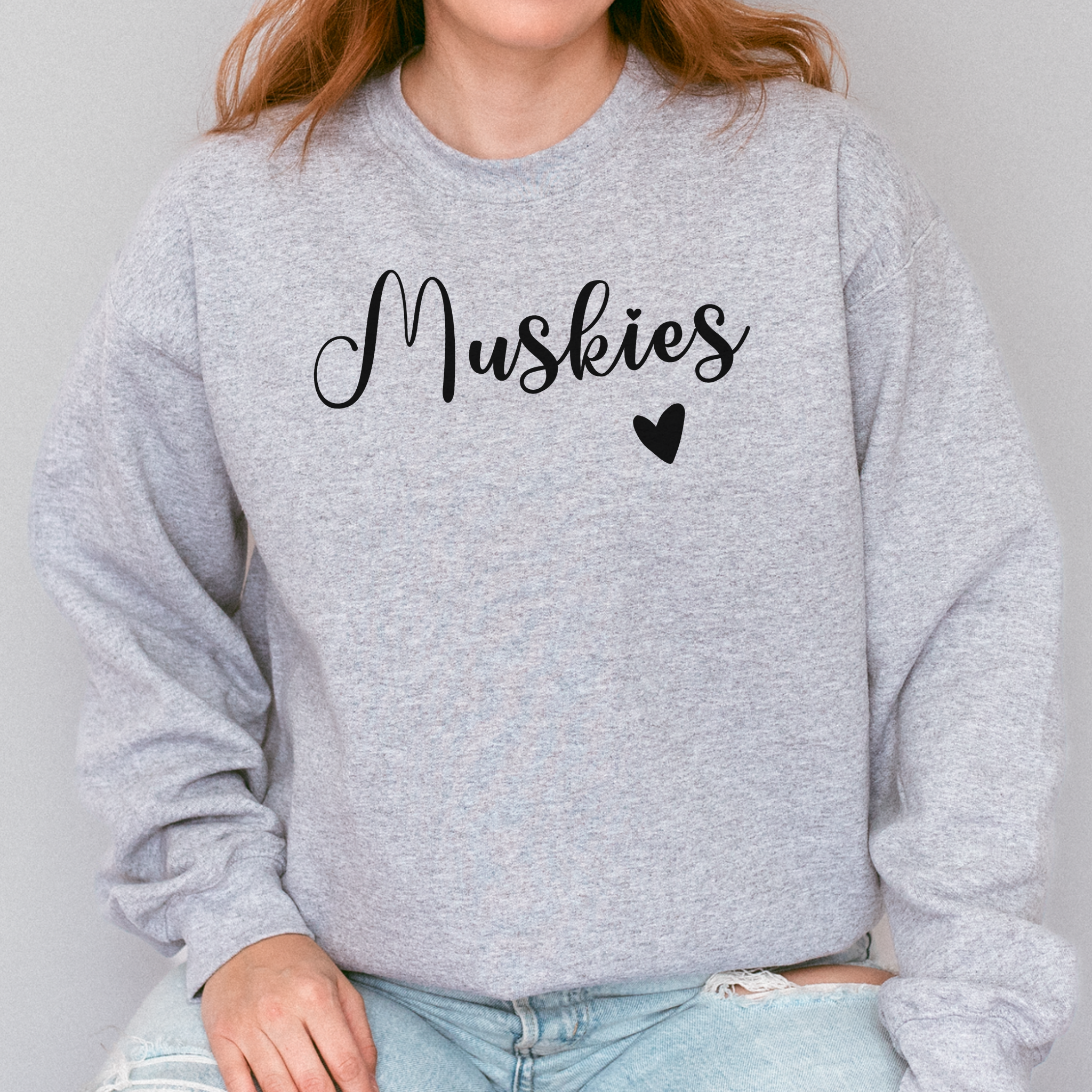 Person wearing a gray sweatshirt with 'Muskies' and a heart graphic on a plain background