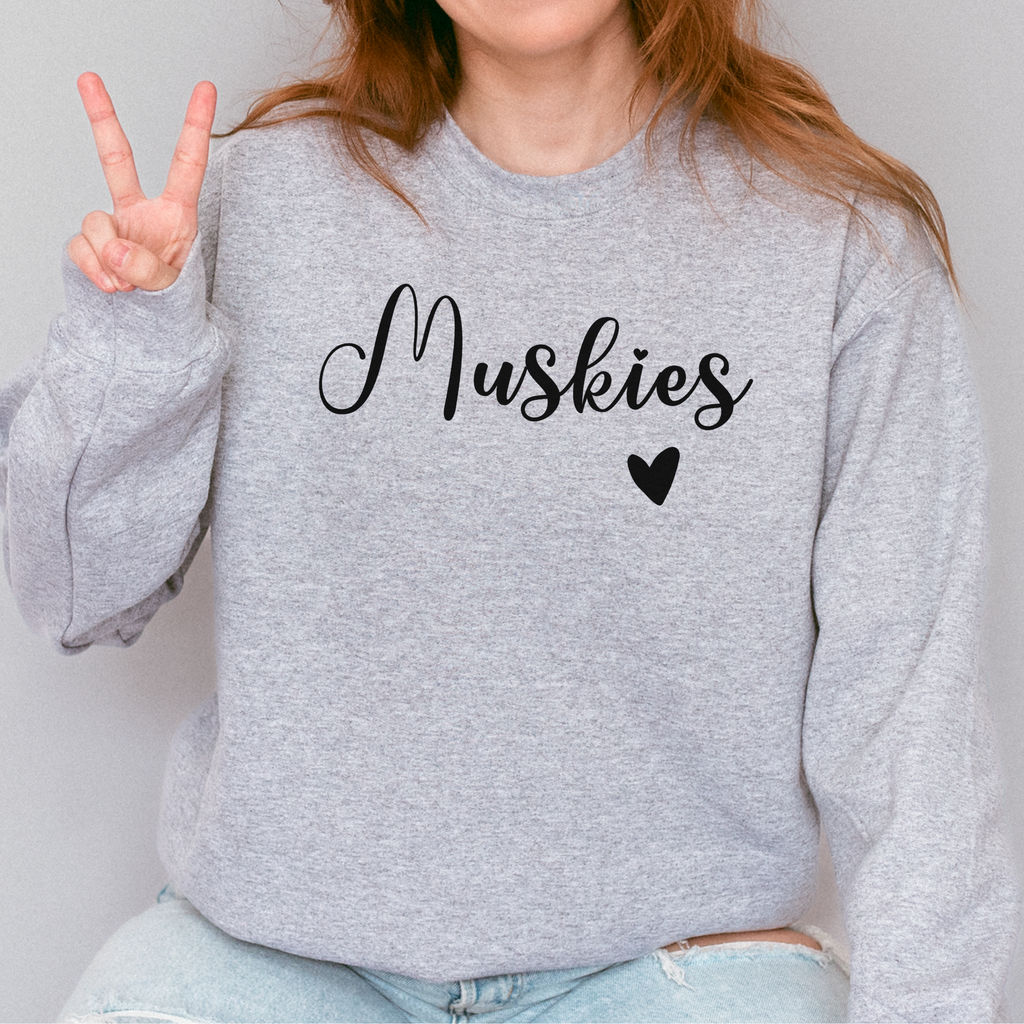 Person wearing a gray sweatshirt with 'Muskies' and a heart design, making a peace sign.
